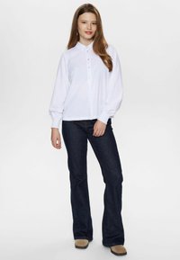 Camicia bianca con maniche lunghe e polsini arricciati, abbinata a jeans a zampa d'elefante blu scuro e calzature beige.
