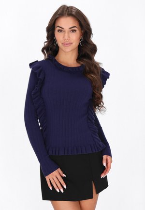 Femme aux longs cheveux bruns ondulés portant un pull bleu marine à volants et une jupe noire fendue devant sur fond blanc.