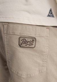 Beige cargoshorts hebben een gestikte zak met een rechthoekig label waarop "Petrok 1980" staat in donkerbruine cursieve letters.