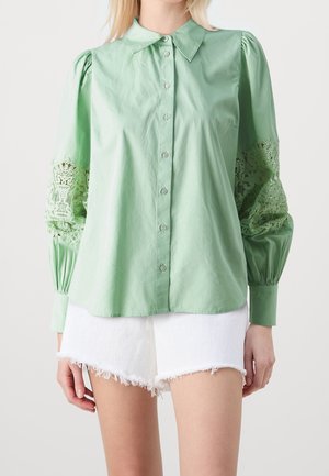 Vrouw draagt een lichtgroene blouse met knopen en kanten mouwen en witte versleten denim shorts, staand tegen een effen achtergrond.