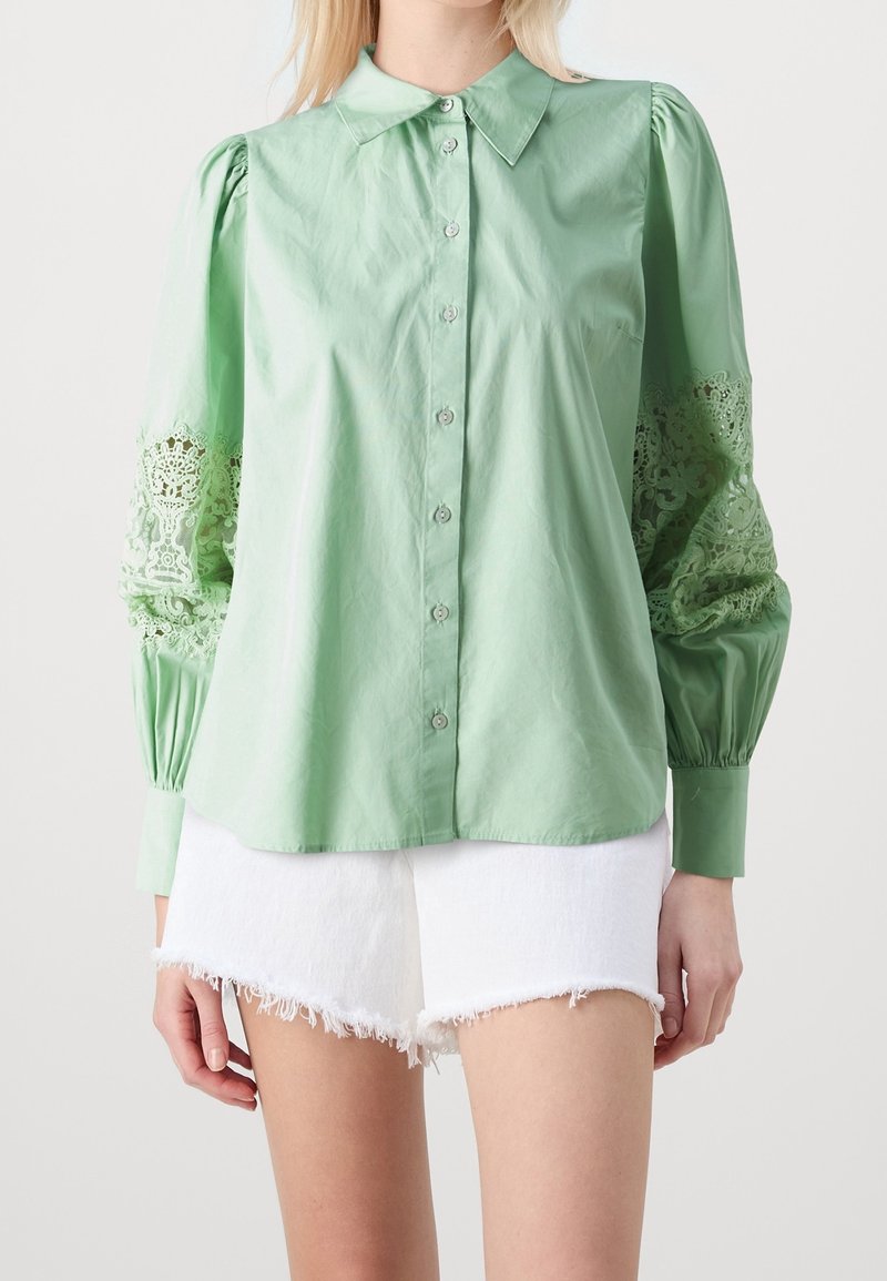 Femme portant un chemisier vert clair boutonné avec des manches en dentelle et un short en jean blanc effiloché, debout devant un fond uni.