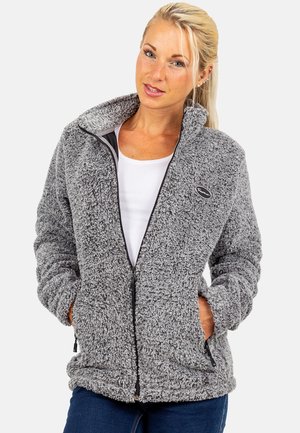 Frau trägt eine graue, flauschige Jacke mit Reißverschluss und die Hände in den Taschen, darunter ein weißes Hemd und blaue Jeans vor einem einfarbigen Hintergrund.