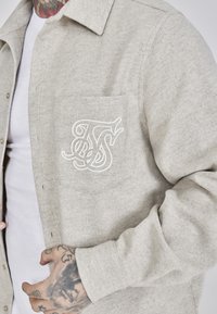 Light gray flannel shirt with a pointed collar, featuring a left chest pocket. Intricate white embroidered logo on the pocket.