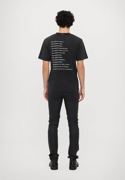 Czarny t-shirt z krótkim rękawem z białym napisem na plecach. Posiada klasyczny okrągły dekolt i lekko dopasowany krój. Noszony z ciemnymi spodniami.