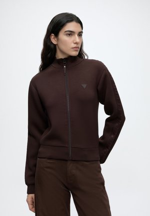 Personne aux longs cheveux foncés portant une veste zippée marron foncé et un pantalon assorti, légèrement tournée vers la gauche avec une expression neutre.