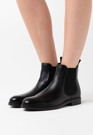 Bottes noires en cuir montantes avec panneaux latéraux élastiques portées sur des jambes nues sur fond blanc.