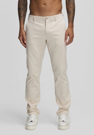 Mężczyzna ubrany w beżowe spodnie o prostych nogawkach i białe trampki, stojący z rękami swobodnie opuszczonymi wzdłuż ciała na jednolitym tle.