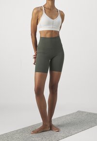 Biały sportowy biustonosz z wysoką linią dekoltu i regulowanymi ramiączkami, zestawiony z oliwkowozielonymi szortami z wysokim stanem. Gładka, elastyczna tkanina.