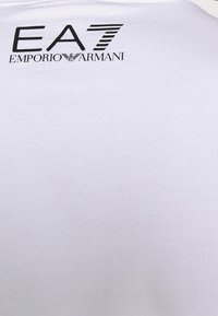 Vit bomull t-shirt med en svart logotyp med texten "EA7" och "EMPORIO ARMANI" på den övre delen av ryggen. Slät textur med en klassisk passform.