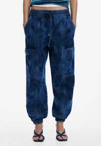 Pantalon cargo en denim bleu foncé avec une taille élastique, des chevilles retroussées et de grandes poches latérales. Caractérisé par une texture douce et un design non structuré.