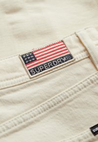 Tela beige con un acabado texturizado, que presenta un parche rectangular con una bandera americana y "SUPERDRY" bordado en azul marino.