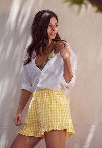 Weißes Leinenhemd, gelbe und weiße Karo-Shorts mit Rüschensaum, grünes Strickoberteil darunter, natürliche Umgebung mit sanften Lichtschatten.