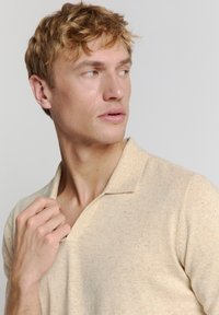 Lichtbeige gebreid poloshirt met korte mouwen, met een zachte textuur en een ingesneden kraag. Subtiele kleurvlekken zijn zichtbaar.