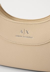 Borsa in pelle beige testurizzata con forma curvilinea e cuciture sottili. Presenta un logo argentato "A/X" e il testo "ARMANI EXCHANGE".