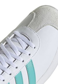 Närbild av en vit sneakersko med vita snören, tealränder på sidan och en texturerad ljusgrå mockatå.