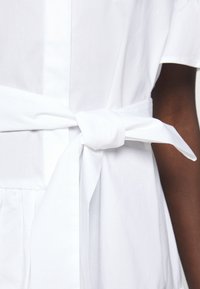 Gros plan d’une personne à la peau foncée portant une robe blanche avec une large ceinture en tissu nouée en nœud à la taille.