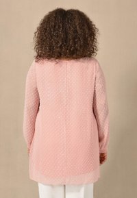 Blusa rosa trasparente con pois testurizzati, maniche lunghe e vestibilità morbida. Presenta un taglio dritto e un collo rotondo.