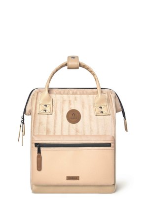 Beige quilted backpack with textured leather accents, a front zip pocket, top handle, and logo patch. Smooth finish with zippers and snaps.