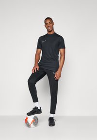 T-shirt et pantalon de sport noirs à coupe ajustée. Le t-shirt présente un logo Nike blanc ; le modèle se tient sur un ballon de soccer. Baskets noires et chaussettes blanches.