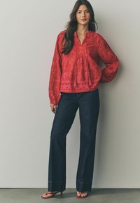 Blusa rossa, lavorata, con scollo a V e maniche lunghe a sbuffo, abbinata a jeans a gamba larga di colore scuro e sandali con cinturini. Tra i dettagli decorativi spiccano le nappine.