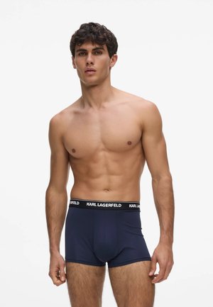 Modèle masculin aux cheveux courts et bouclés portant un boxer bleu marine avec une ceinture noire estampillée Karl Lagerfeld sur fond blanc.