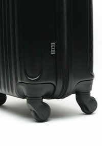 Valigia rigida nera con superficie texture, ruote multidirezionali e zip, con un piccolo etichetta del marchio sul lato.
