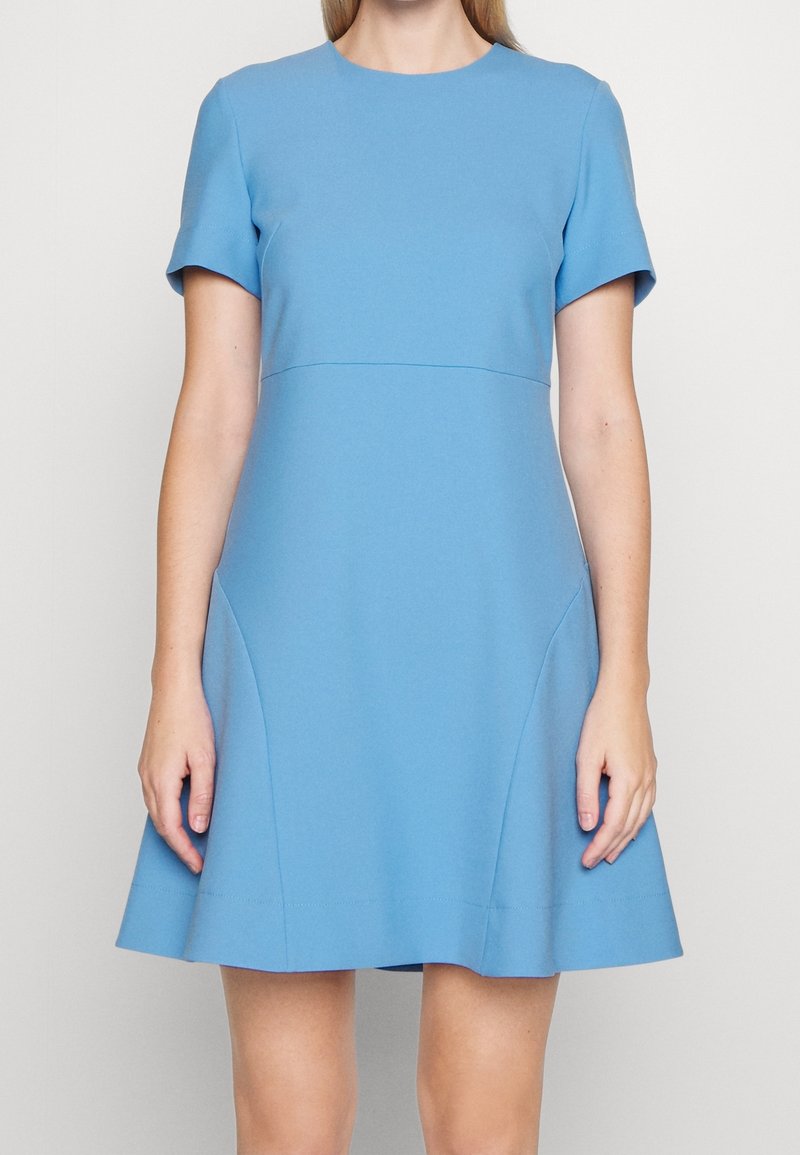 Femme portant une robe bleu clair à manches courtes, longueur genou, avec un corsage ajusté et une jupe évasée, debout devant un fond uni.