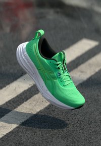 Ténis desportivo verde com parte superior em rede, entressola branca e interior preto. Apresenta detalhes amarelos e superfície texturada para aderência.