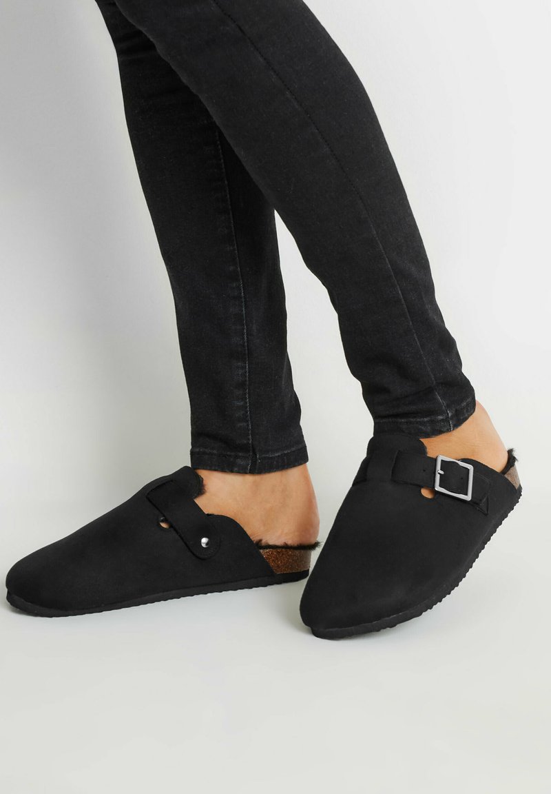 Mules de ante negro con puntera redondeada, detalle de hebilla lateral y plantilla texturizada marrón. Llevados con unos pantalones vaqueros oscuros ceñidos.