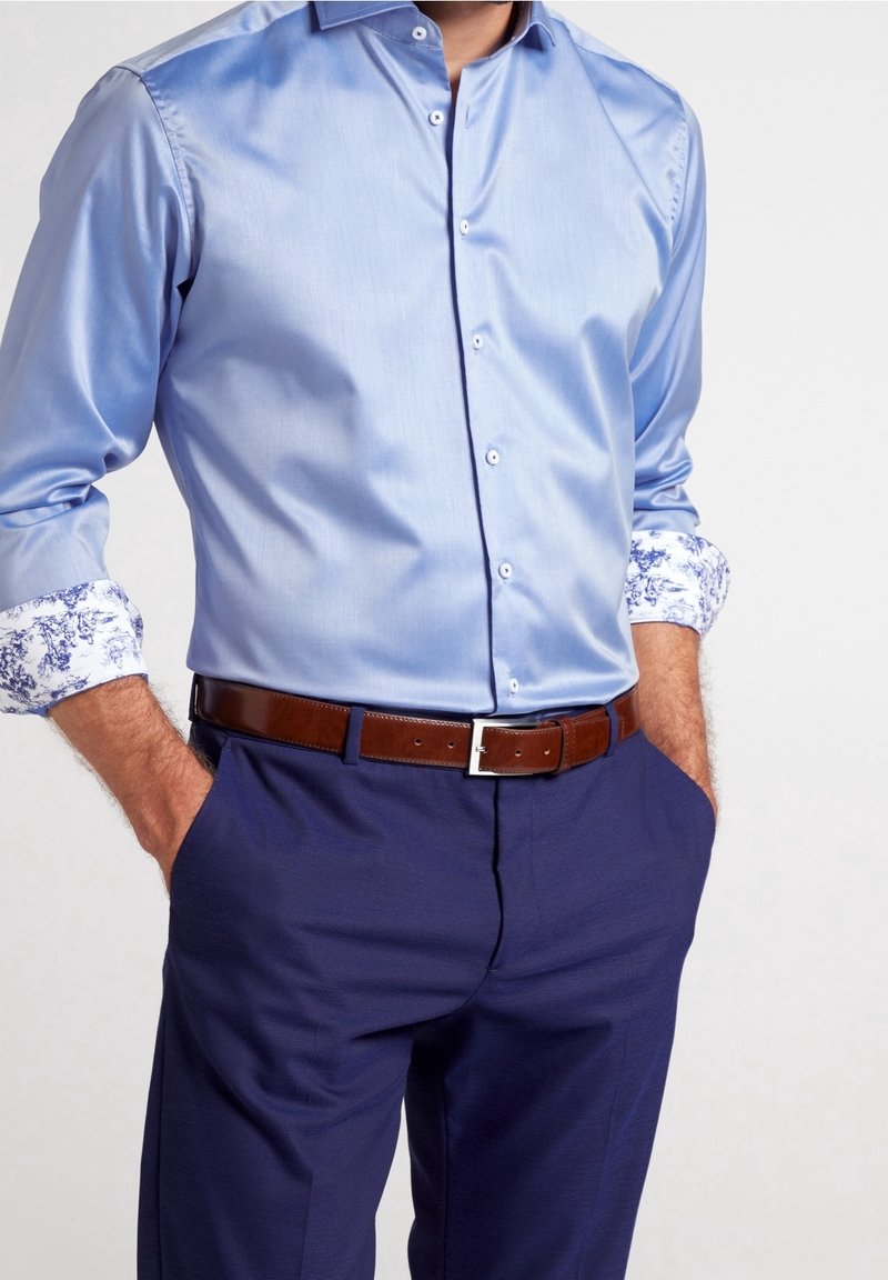 Camicia da uomo satinata di colore azzurro chiaro con dettagli floreali bianchi sui polsini, abbinata a pantaloni blu scuro e una cintura in pelle marrone.