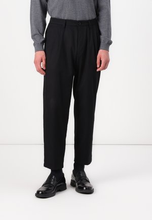 Black pleated trousers made of soft fabric, featuring a high waist, tapered legs, and a button closure. Worn with black loafers.