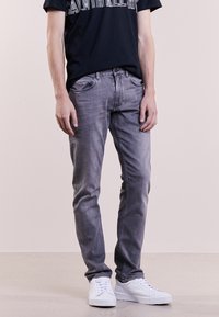 Hombre con jeans ajustados de color gris desgastado, camiseta gráfica negra y zapatillas blancas, de pie contra un fondo claro liso.