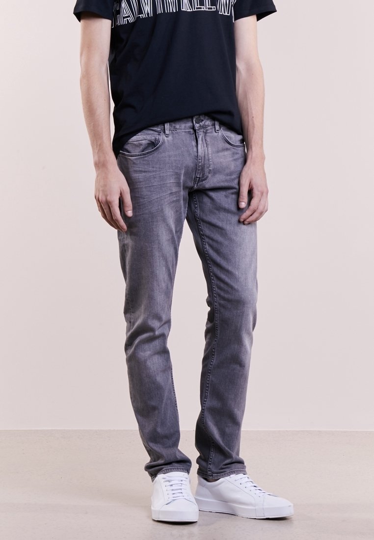 Hombre con jeans ajustados de color gris desgastado, camiseta gráfica negra y zapatillas blancas, de pie contra un fondo claro liso.