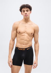 Γυμνόστηθος νεαρός με σγουρά μαλλιά φορεί μαύρα boxer της Nike με πορτοκαλί λογότυπο στη μέση, μπροστά σε απλό φωτεινό φόντο.