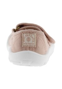 Zapatilla de lona suave en color rosa claro, con suela de goma blanca, que presenta una pequeña etiqueta de marca en la parte trasera y un diseño de tacón redondeado.