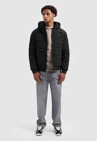 Zwarte pufferjack met capuchon, gewatteerd ontwerp en ritsluiting, gedragen over een beige sweatshirt. Gecombineerd met lichtgrijze jeans en zwarte sneakers.