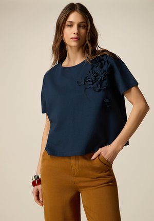 Donna con indosso una maglietta a maniche corte blu navy con applicazioni floreali sulla spalla e pantaloni color senape, in posa con una mano in tasca.