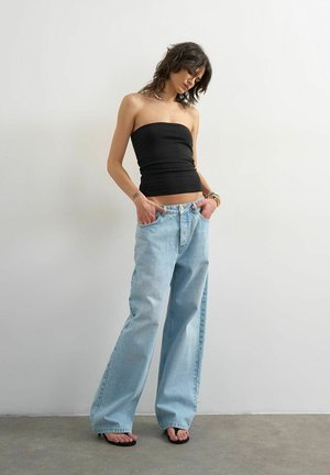 Femme portant un haut noir sans bretelles, un jean large bleu clair et des sandales noires, debout contre un mur blanc uni, les mains dans les poches.