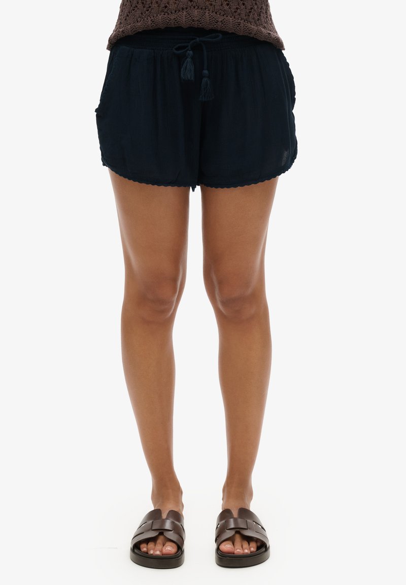 Shorts bleu foncé avec une coupe ample, ceinture à cordon de serrage et ourlet festonné. Portés avec des sandales marron, laissant voir les orteils.