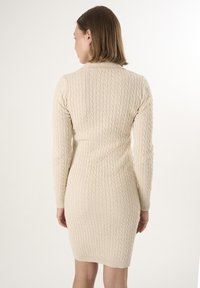 Abito lavorato a maglia beige con motivi a treccia, maniche lunghe, silhouette aderente e polsini e orlo a coste, visto da dietro.