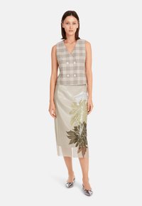 Kariertes, doppelt geknöpftes Weste in Beige und Grau, kombiniert mit einem halbtransparenten, beige Rock mit floralen Mustern in Grün und Gold.