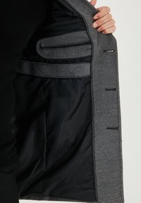 Sivi volneni plašč s črnim podlogom, notranjimi žepi, dekorativnim šivanjem in krojenim dizajnom. Gladek tekstura in čist zaključek.