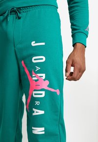 Turkosa joggbyxor i mjukt tyg, med vit text "JORDAN" och en rosa Air Jordan-silhuettgrafik på vänster ben.