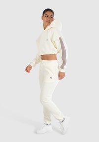 Sudadera corta de marfil con capucha y acentos grises en las mangas, combinada con joggers de marfil que tienen bolsillos y un logo, usados con zapatillas blancas.