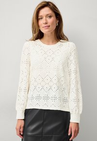 Blouse en dentelle blanche avec un motif floral, encolure ronde et manches longues. Design texturé avec des détails en ajouré sur l'ensemble.