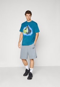 Ung mand står op, iført en blå grafisk T-shirt, grå Nike-shorts, hvide Nike-sokker og sorte sneakers mod en ensfarvet baggrund.