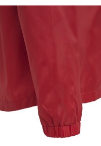 Giacca impermeabile rossa con finitura liscia e lucida. Presenta polsini elasticizzati e una vestibilità rilassata, adatta per diverse condizioni meteorologiche.