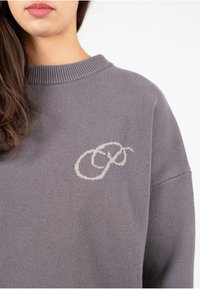 Pull en maille gris avec un col rond et des détails en côtes. Présente un design brodé de couleur claire sur la poitrine, texture douce.