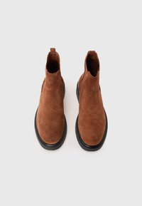Botine din suede maro, cu o textură netedă, având o talpă din cauciuc negru și un sistem de prindere la spate pentru o îmbrăcare ușoară.