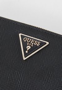 Materiale nero testurizzato con un logo a triangolo accentuato in oro che presenta "GUESS USA" e un punto interrogativo su uno sfondo nero lucido.